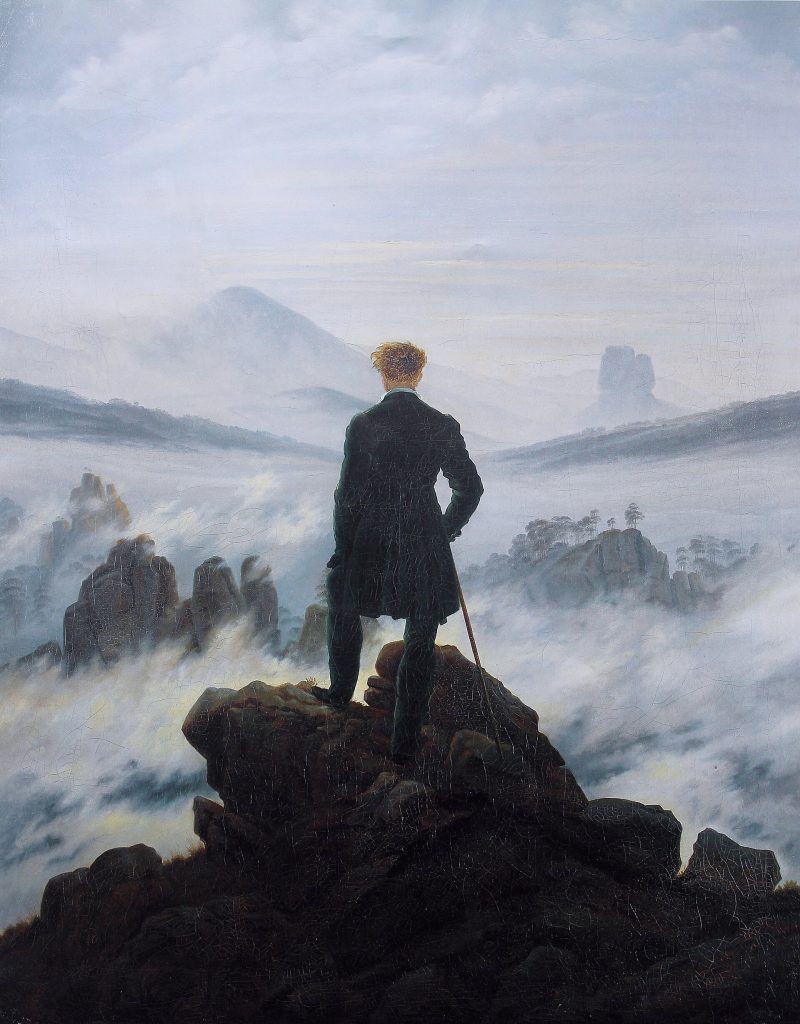Man with a walking stick on a rocky summit gazing over fog-covered mountains